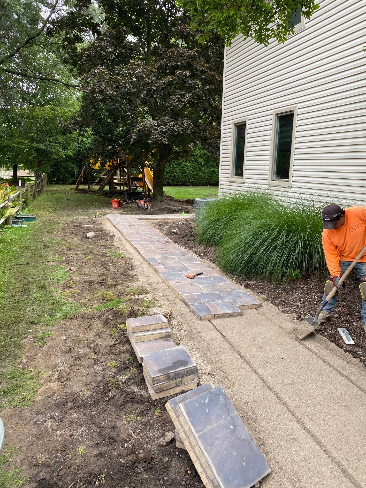 Worker shoveling beside a house walkway during landscaping, with stone pavers and trees nearby