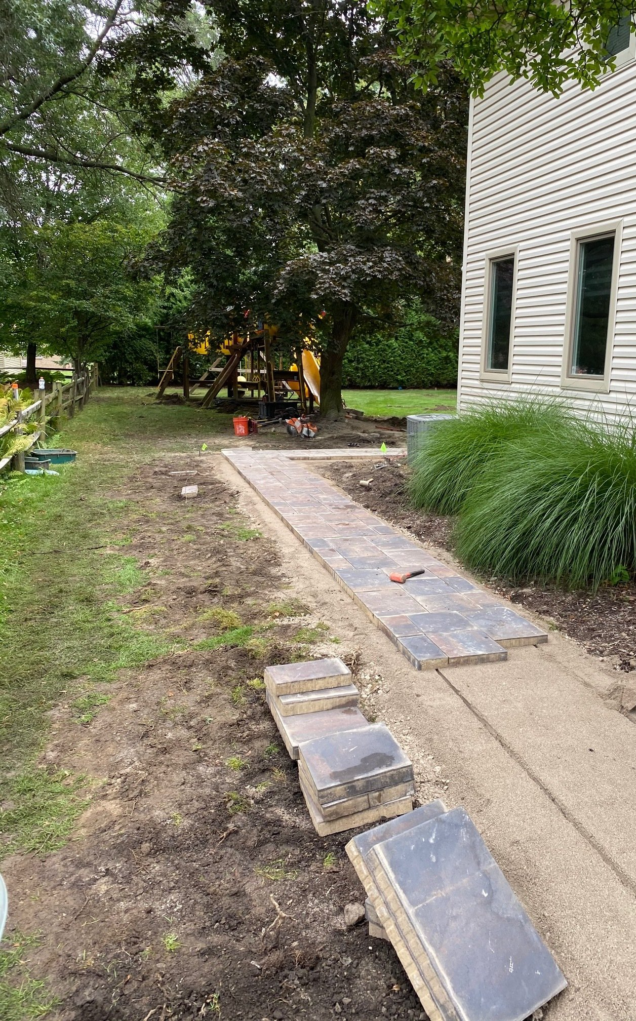Backyard path beside a house with stepping stones, gravel, grass, and trees leading to a shaded patio area
