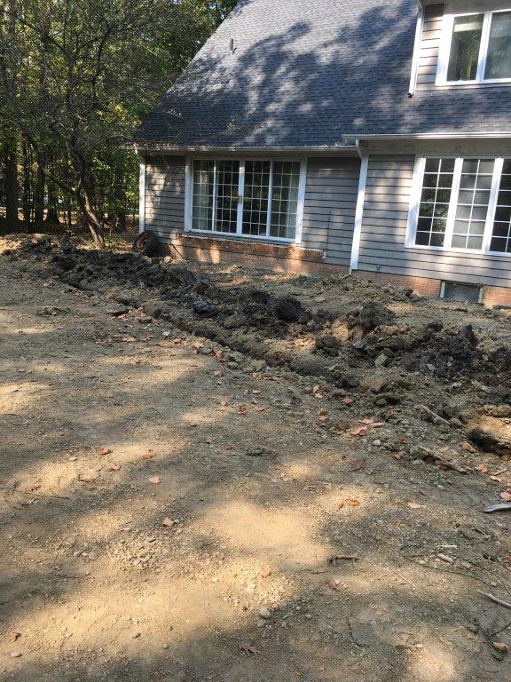 Dirt trench dug beside a gray house with white-trimmed windows in a wooded yard