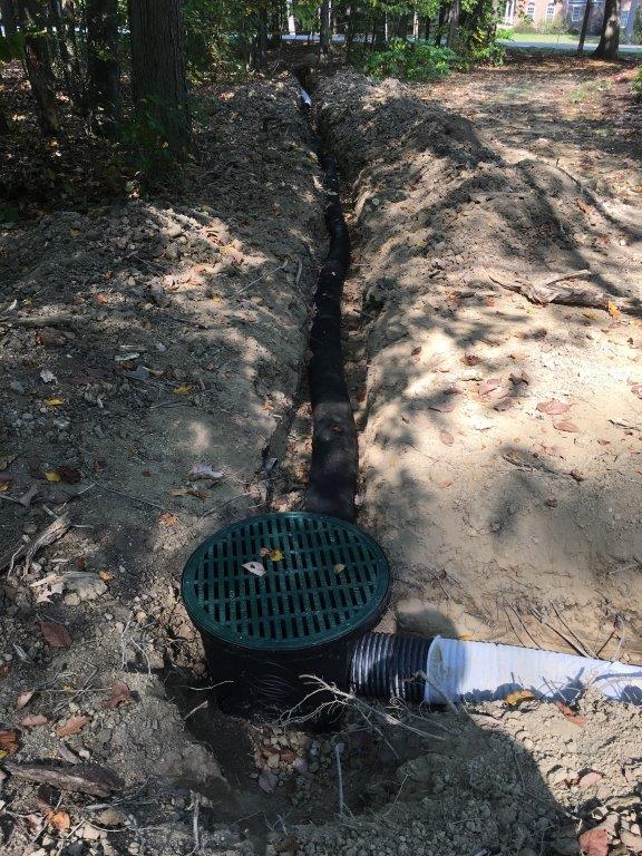 A roadside drainage ditch with a round green storm drain grate beside a white pipe.