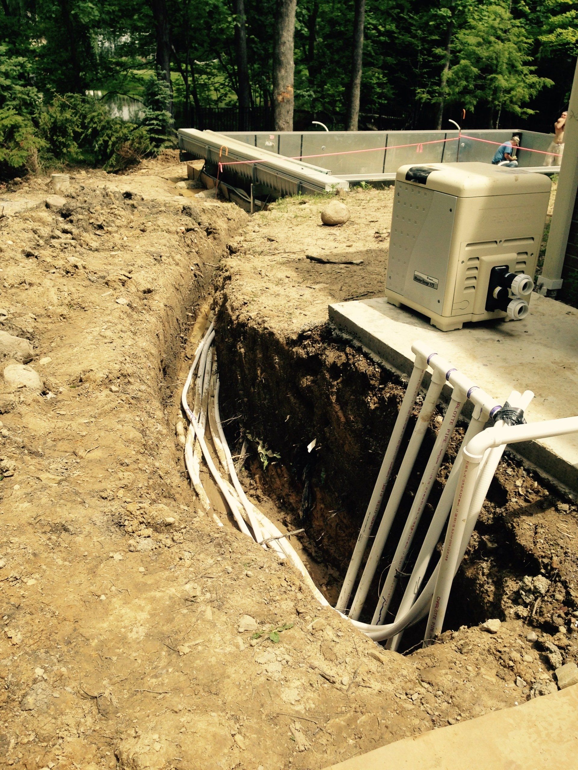Open trench beside a road, with exposed pipes and a utility box in a wooded area