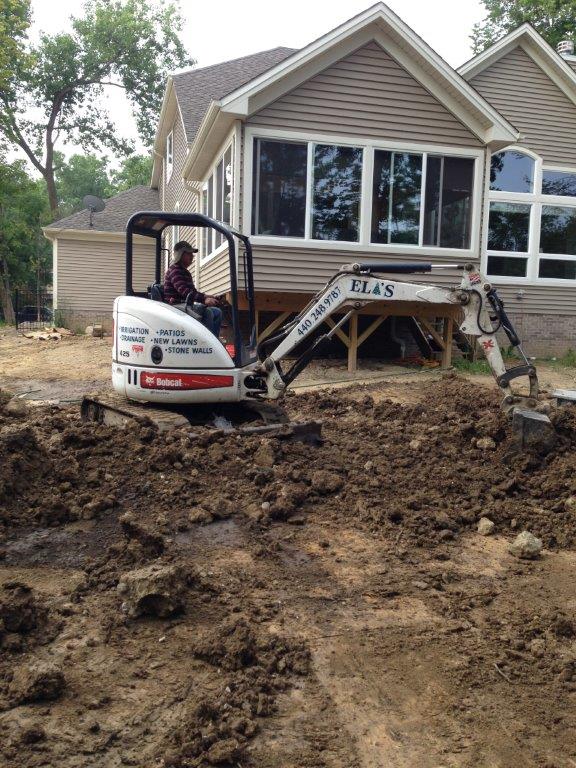 Mini excavator grading muddy soil in front of a house with a covered porch and large windows
