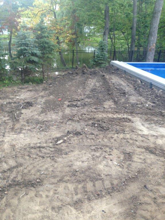 Dirt mound beside a blue above-ground pool in a wooded backyard with a chain-link fence