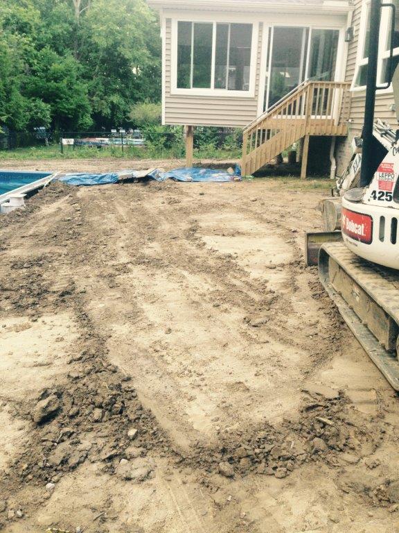 Dirt driveway under construction beside a house, with a skid steer and a blue tarp in the yard.