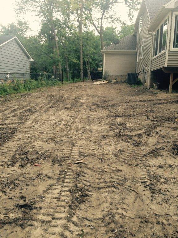Mud-covered backyard beside a house, with tire tracks and trees in the background