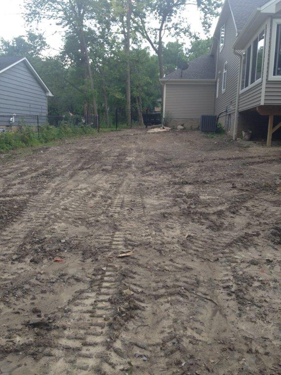 Mud-filled backyard beside a house, with tire tracks and trees in the background.