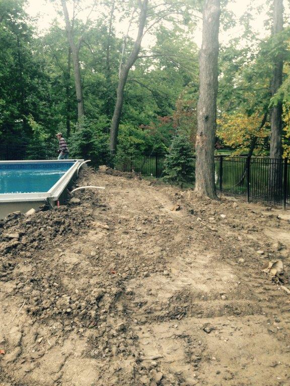 Dirt-filled backyard beside a swimming pool, with trees and a black fence in the background.