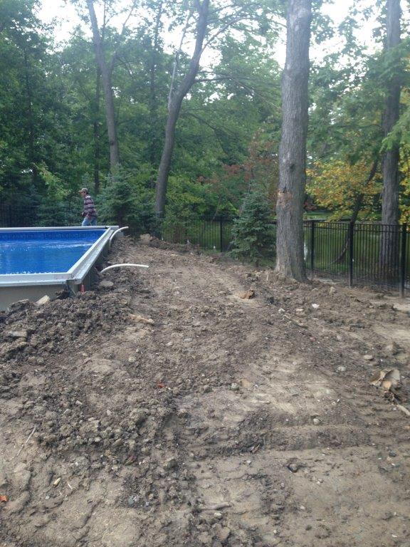A muddy yard beside an above-ground pool, with trees and a fence in the background.