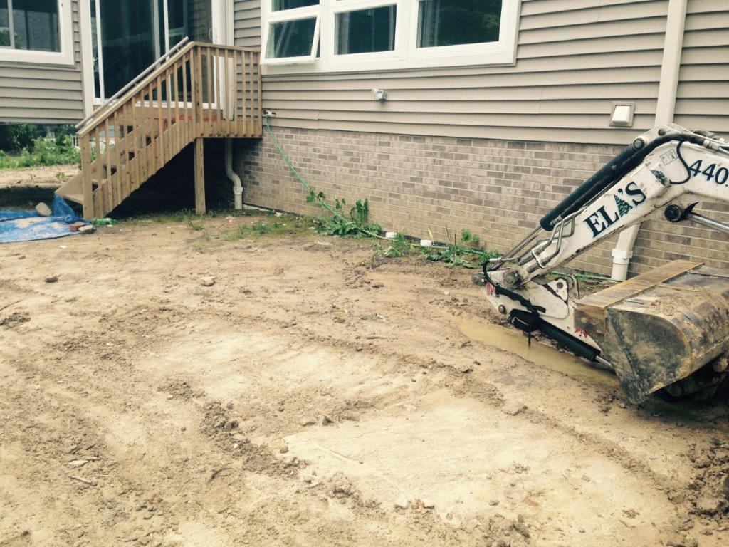 Backyard under construction beside a house, with a compact excavator and freshly graded dirt.