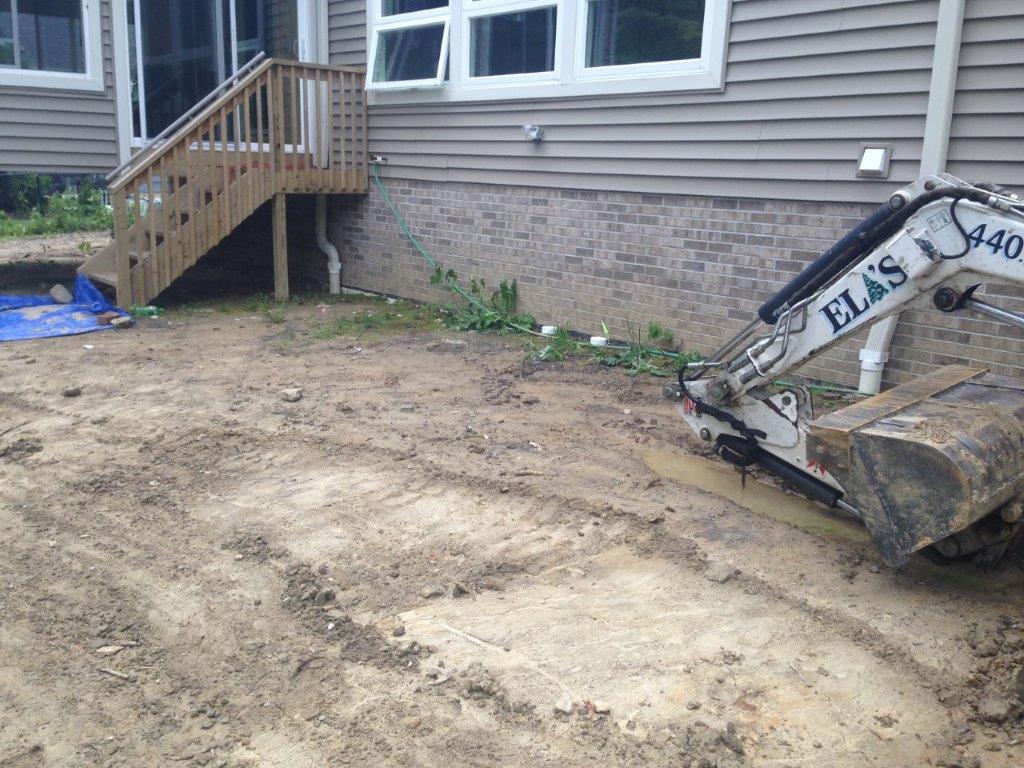 Backyard with dirt cleared near a house, wooden steps, and a small excavator at right.