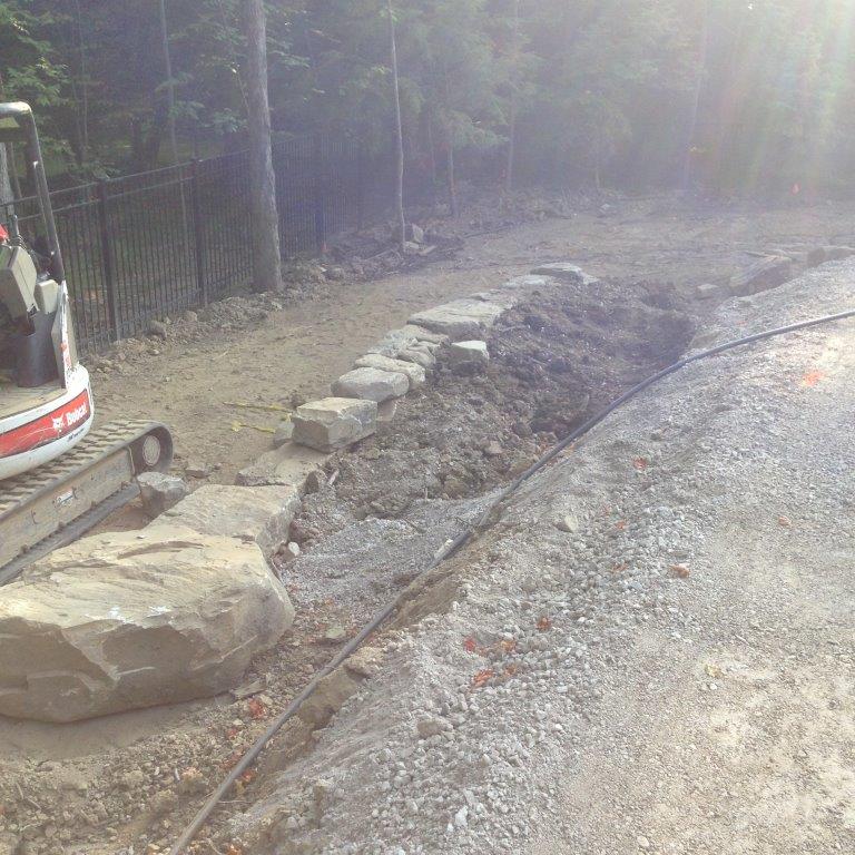 Roadside trench with large rocks and a curb beside a paved street in bright sunlight