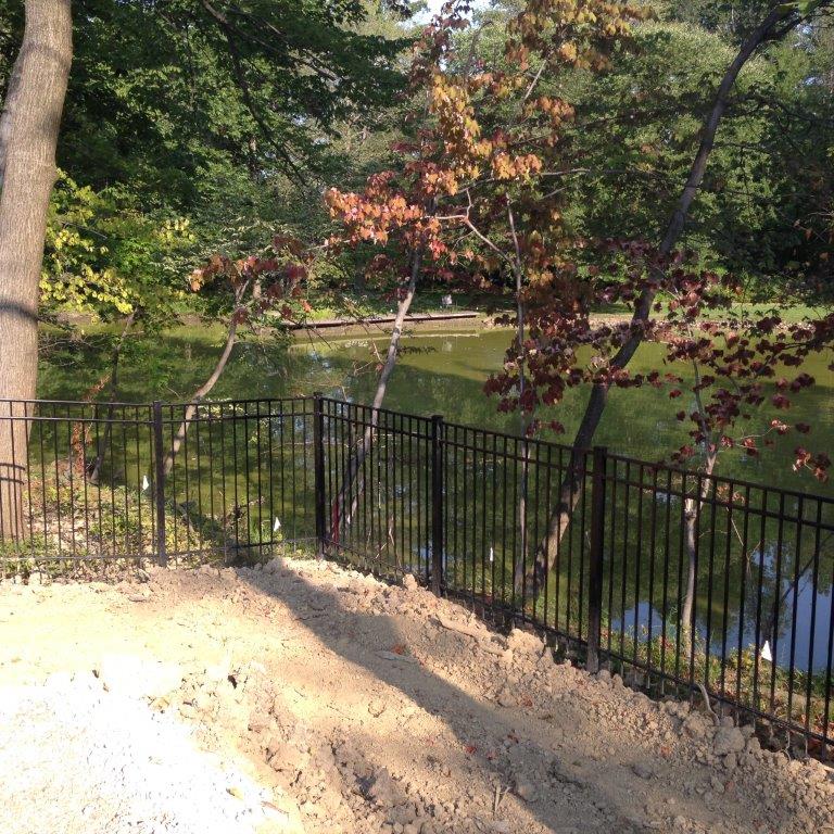 Wooded park with black fence, sunlit path, and a small stream or pond in the background