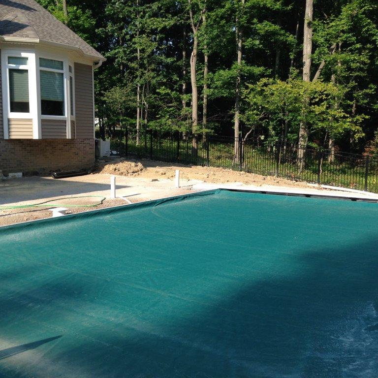 Backyard swimming pool beside a house and wooded fence line on a sunny day