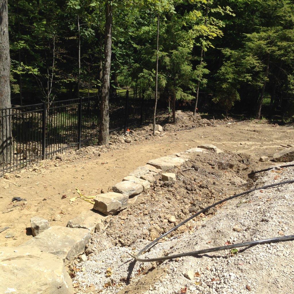 A dirt path beside a stone border and black fence, with trees and brush in the background.