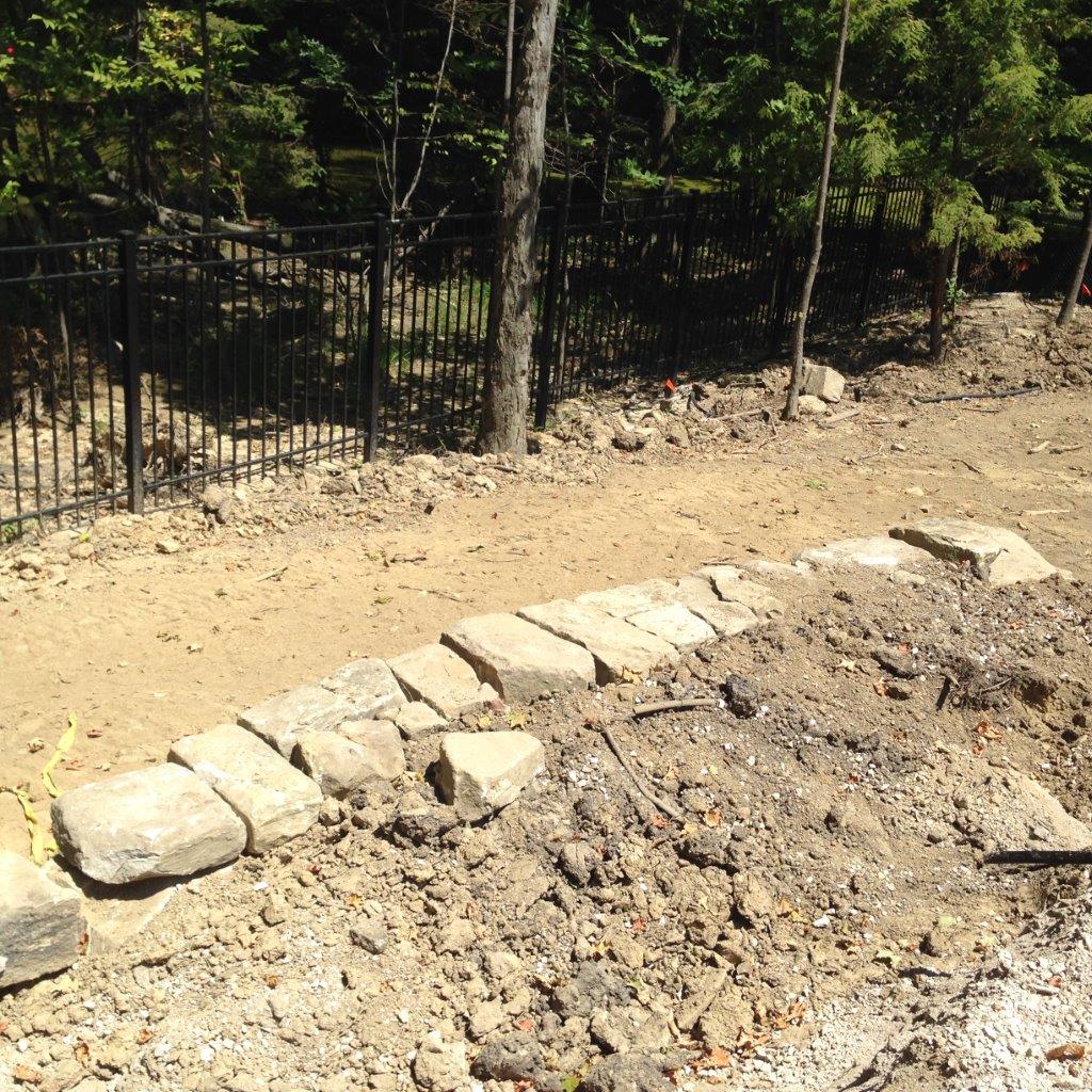 Stone-lined dirt path beside a black metal fence and trees in bright daylight