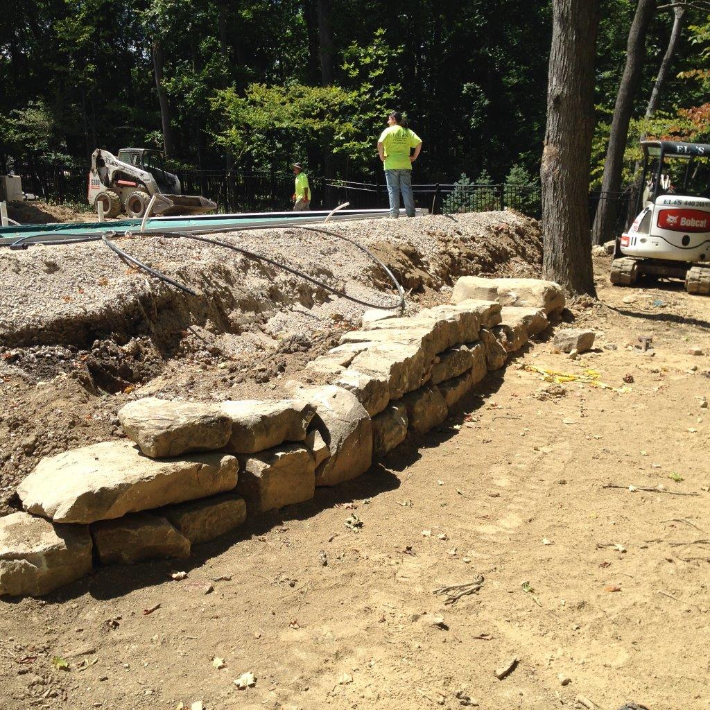 Stone retaining wall under construction beside a dirt path, with workers and a small vehicle nearby.