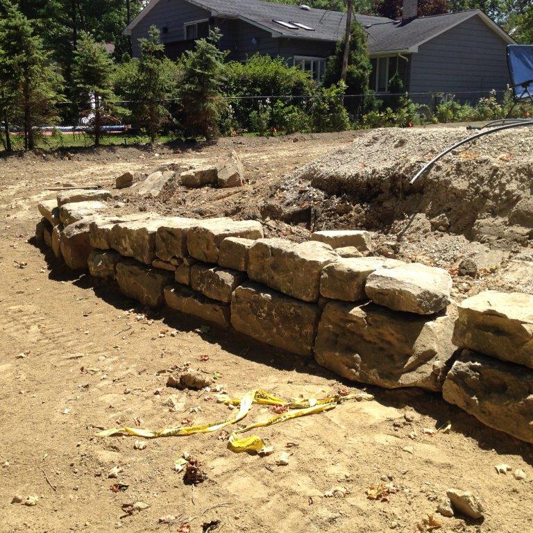 Stone retaining wall under construction on a dirt slope in a sunny yard, with houses and trees in the background