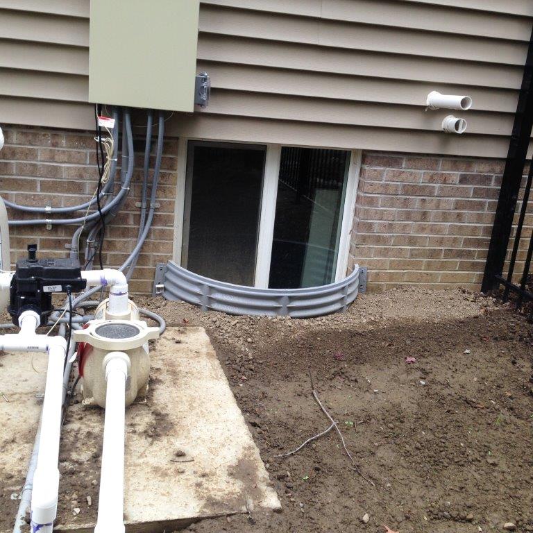 Basement window well beside utility pipes and dirt trench along a house foundation