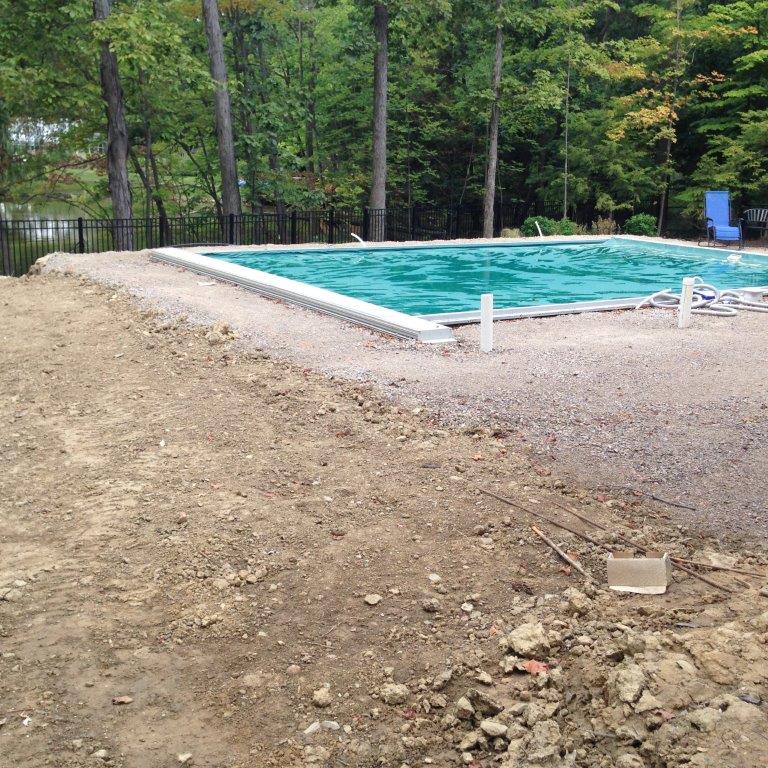 Outdoor swimming pool beside a dirt lot, surrounded by trees and gravel.