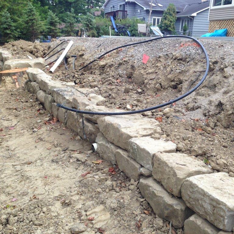 Stone retaining wall under construction on a dirt slope, with a black hose looped over the embankment.
