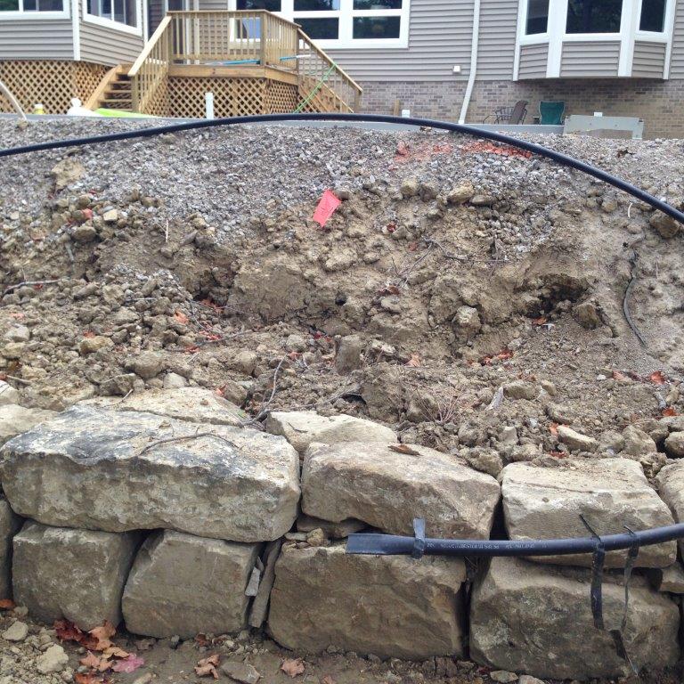 Collapsed stone retaining wall with exposed dirt slope and a black hose in a backyard.