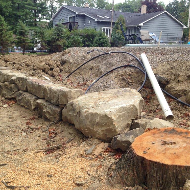 Stone retaining wall and large rocks in a yard with a house in the background, beside a cut tree stump and pipes.