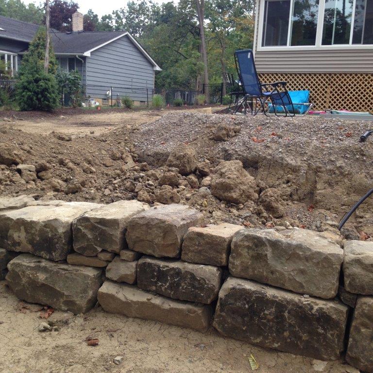 Stone retaining wall beside a large dirt excavation in a backyard, with a house and deck in the background