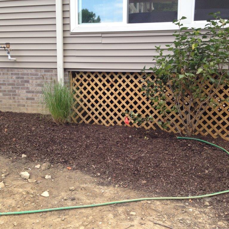 House exterior with tan siding, lattice skirting, mulch bed, and a green garden hose in front of the window