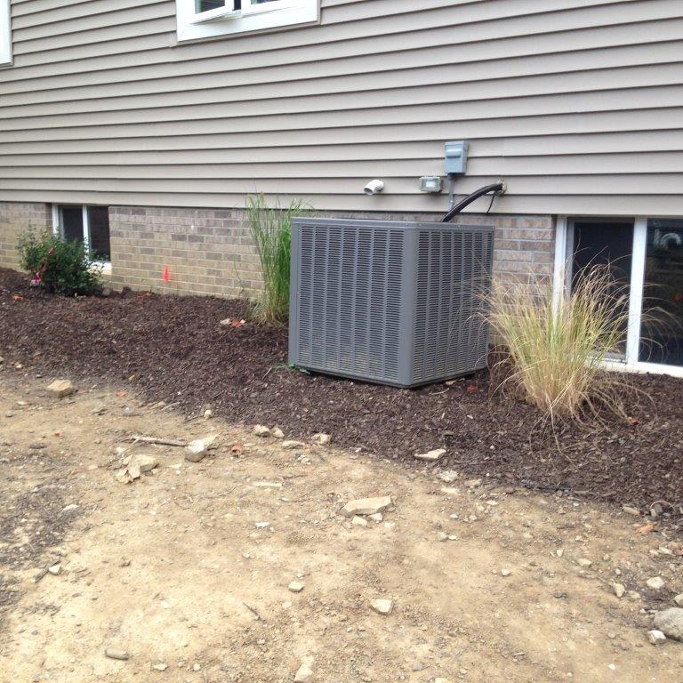 Gray HVAC unit beside a house wall, with mulch bed and a dirt driveway in front.