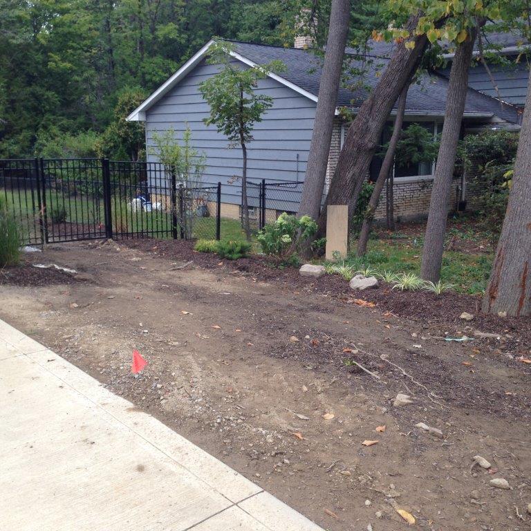 Dirt path beside a concrete driveway, black fence, and gray house in a wooded yard