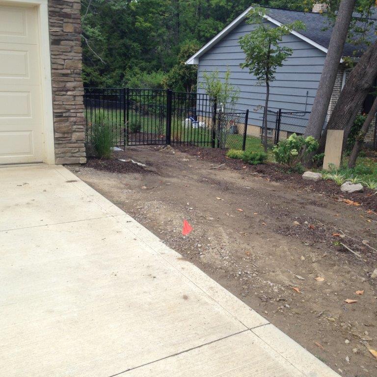 Driveway beside a garage with a dirt side yard, black fence, and small gray shed in the background.