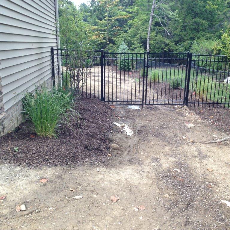 Black metal fence gate beside a house, with a dirt path and garden bed