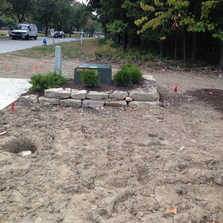 A small rock garden with shrubs and a planter beside a dirt area near a road and trees.