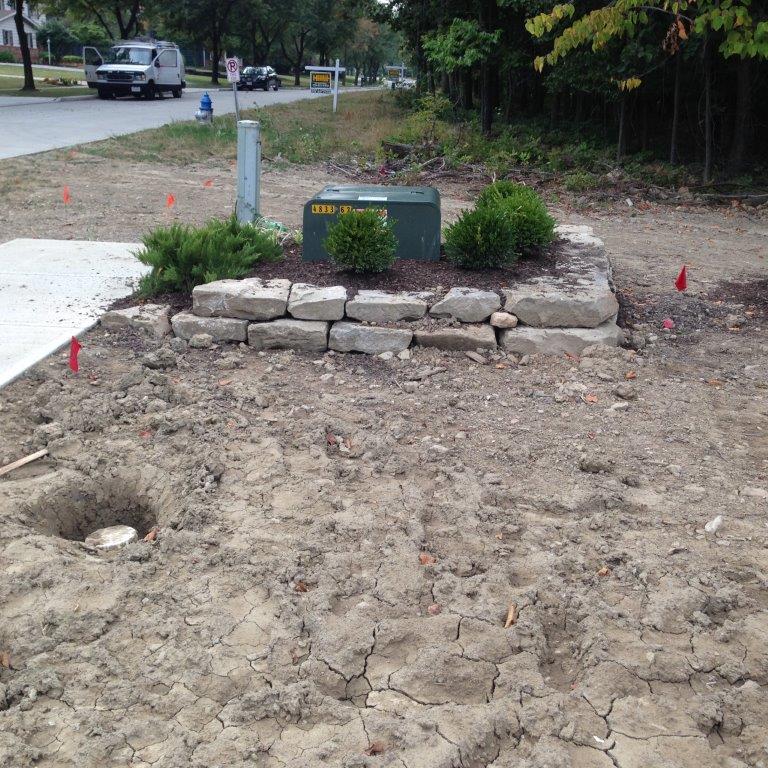 Stone landscaping around a utility box at a dirt roadside corner, with small shrubs and a sidewalk.