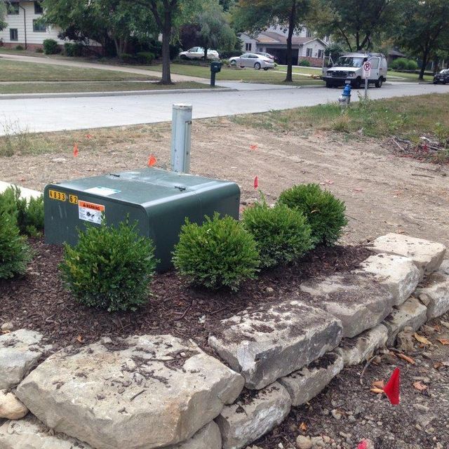 Utility box in a landscaped yard beside a street, bordered by rocks and small shrubs.
