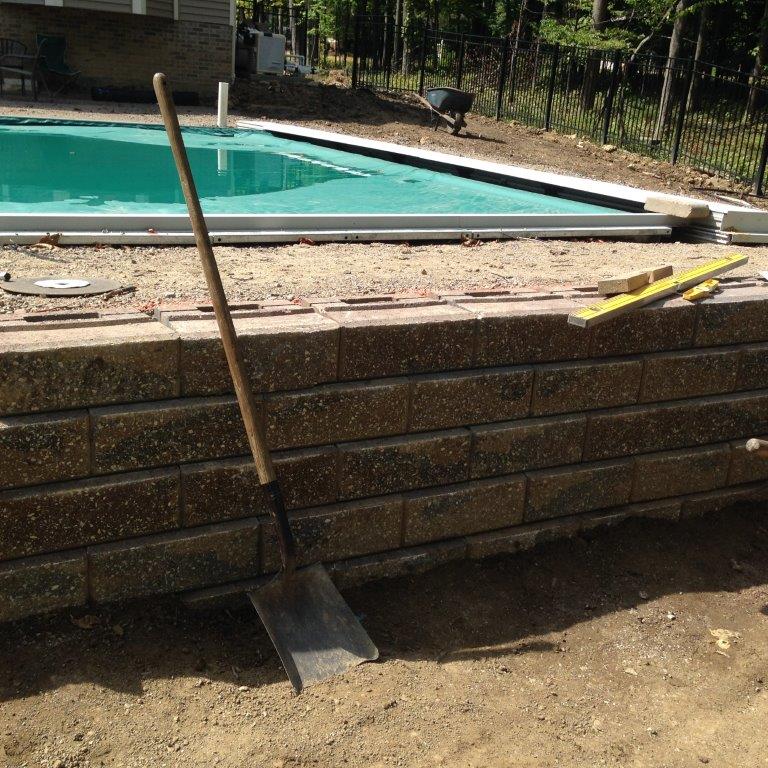 Backyard pool under construction with a shovel leaning against a stone retaining wall.