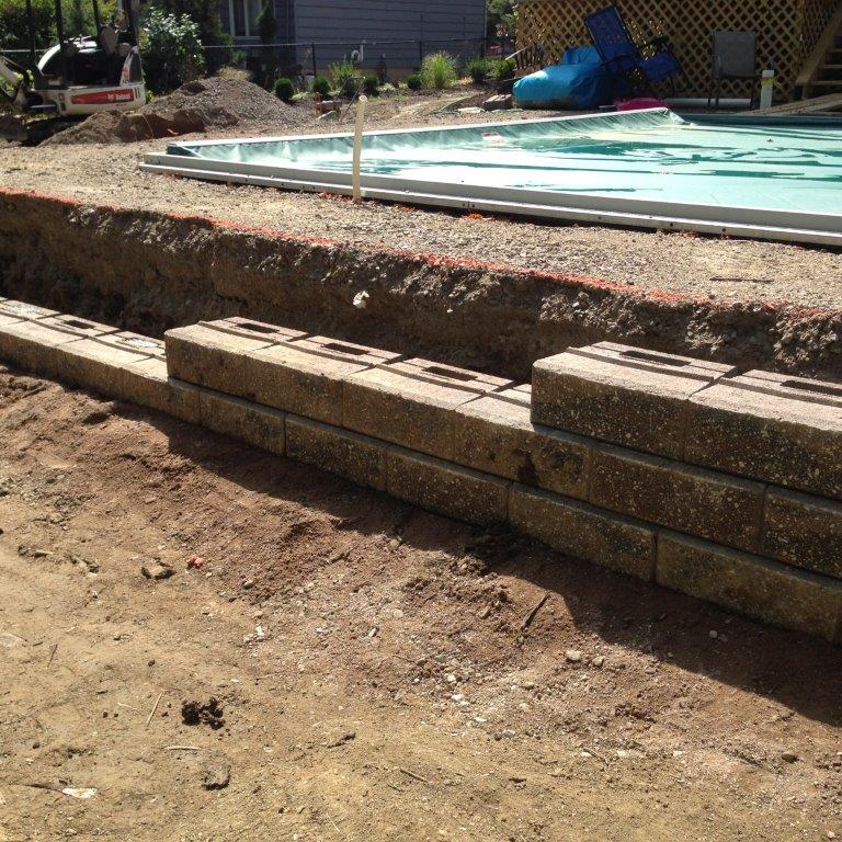 Partially dug backyard pool with stacked concrete retaining wall blocks and dirt foreground
