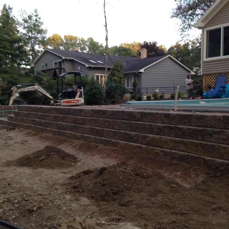 Backyard construction site with excavator, tiered retaining wall, and unfinished dirt yard beside homes