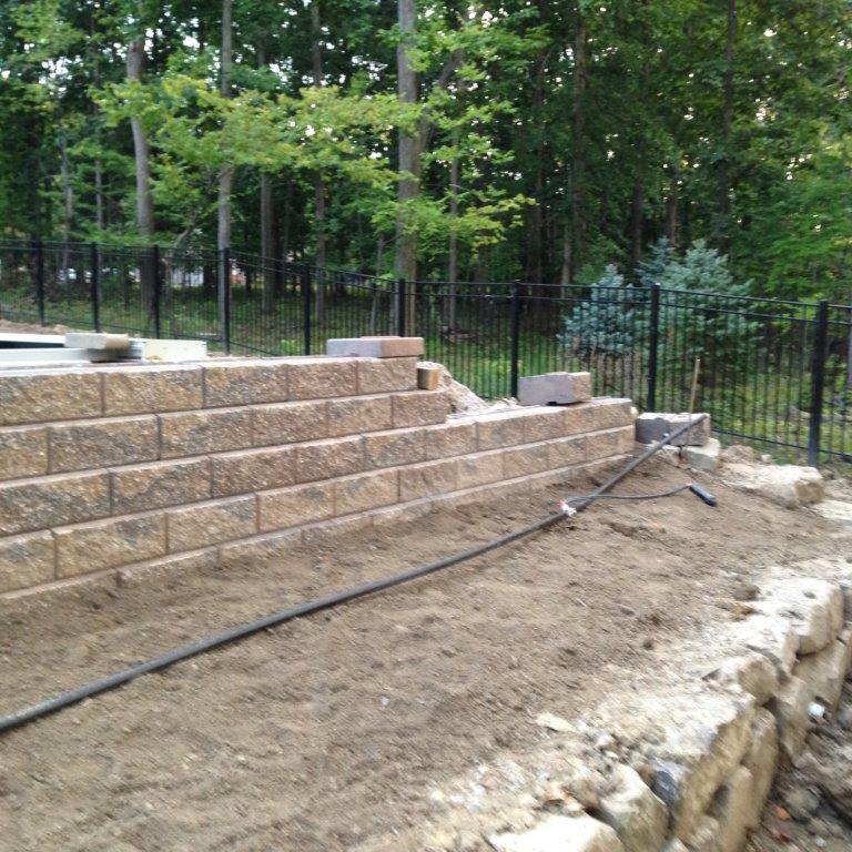 Stone retaining wall under construction beside a dirt yard, with black fence and trees in the background