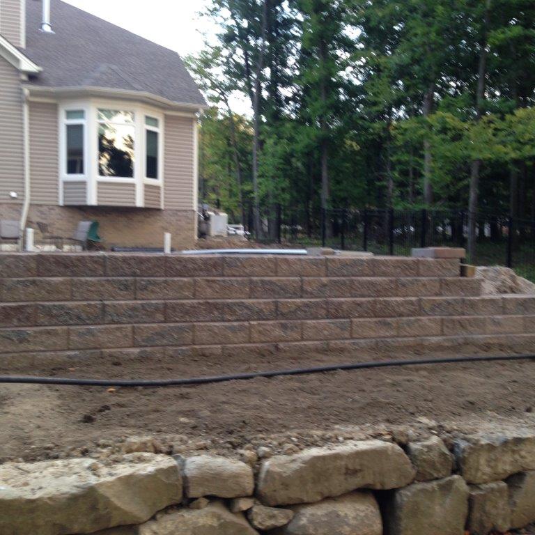 Stone terraced retaining wall beside a tan house, with trees and a rough dirt yard in front.