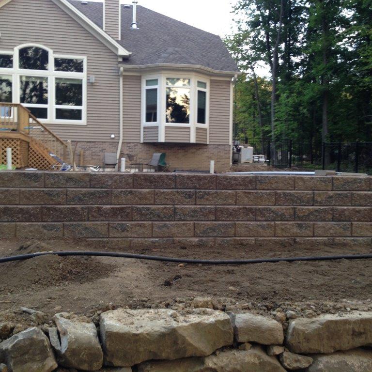 Backyard stone retaining walls and a beige house with a large window and wooded backdrop