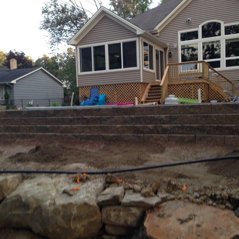 Backyard home with stone retaining wall, steps, and deck beside a large windowed house