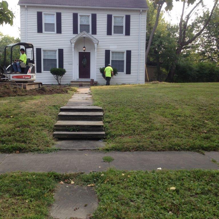 Two workers building stone steps up a grassy slope to a white house with black shutters