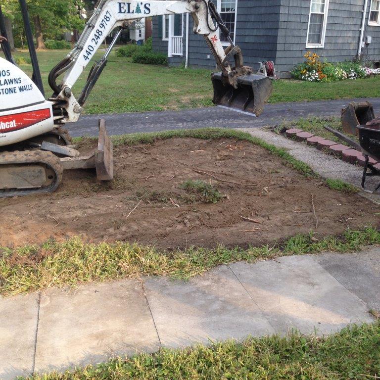 Excavator arm digging up a patch of lawn beside a sidewalk in a residential yard.