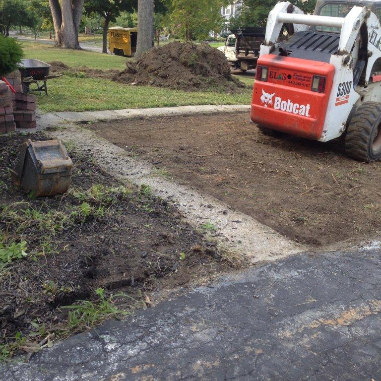 Bobcat skid steer grading a yard beside a driveway, with dirt piles and a tree in the background