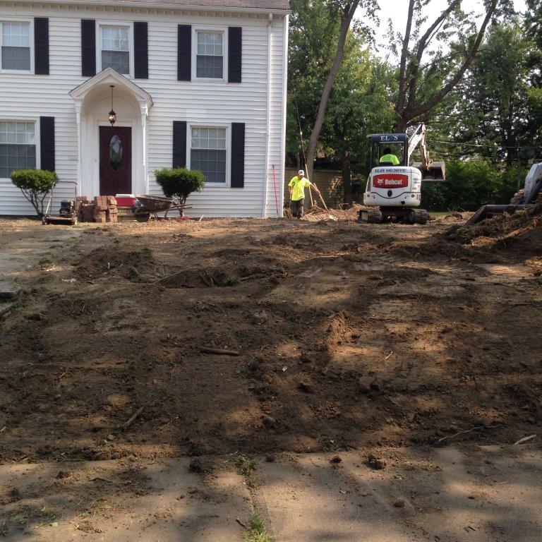 Workers and a skid steer clearing dirt in front of a white house with black shutters