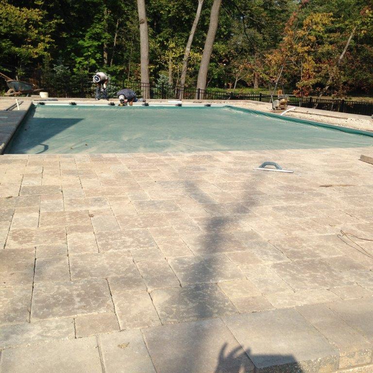 Empty backyard swimming pool with light blue water and stone patio, surrounded by trees.