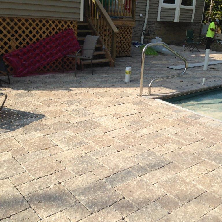 Poolside patio with beige pavers, steps, metal handrail, and a worker in a neon vest at right.