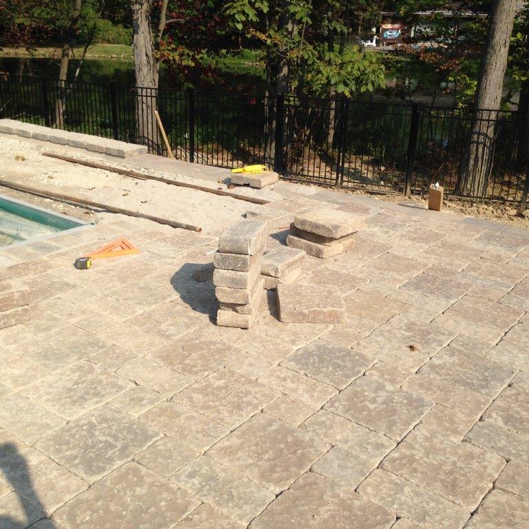 Stone patio beside a pool with a small stacked-stone stool and trees along a fence