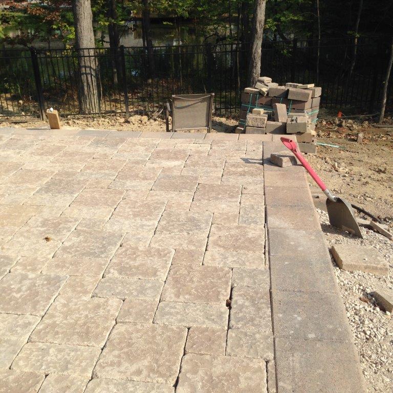 Stone patio under construction beside a fence, with pavers, dirt, and a red-handled shovel.